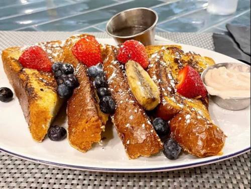 A plate of French toast topped with strawberries, blueberries, and banana, served with syrup and a side of cream.