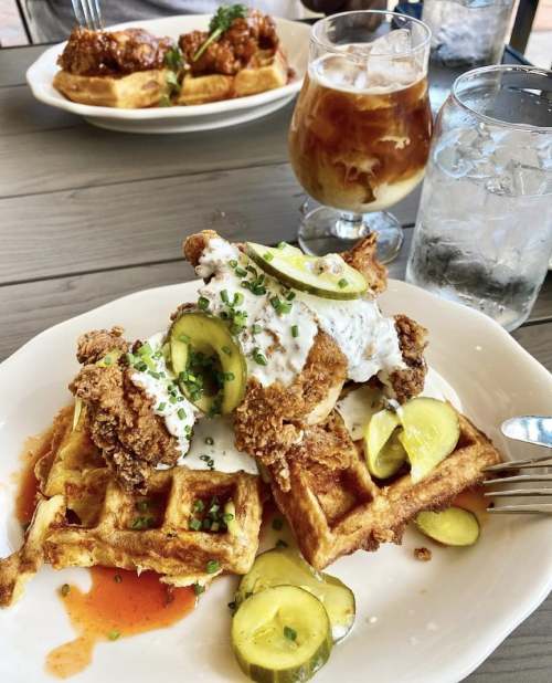A plate of chicken and waffles topped with pickles and drizzled with sauce, alongside a glass of iced coffee.