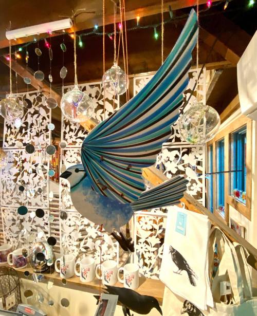 A colorful bird sculpture with outstretched wings hangs in a cozy shop, surrounded by decorative items and lights.
