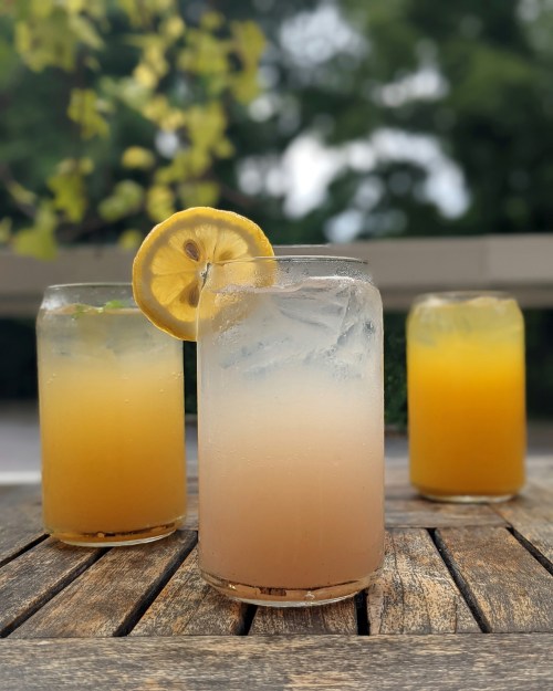 Three refreshing drinks on a wooden table: one with a lemon slice, and two in shades of yellow and orange.