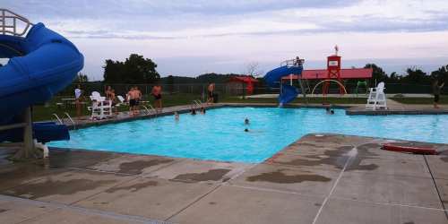 A community pool with a blue slide, children swimming, and adults relaxing nearby under a cloudy sky.