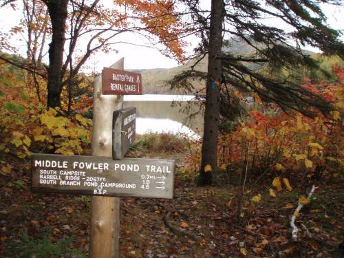 Trail signpost with directions to Middle Fowler Pond Trail, surrounded by autumn foliage and a view of a pond.