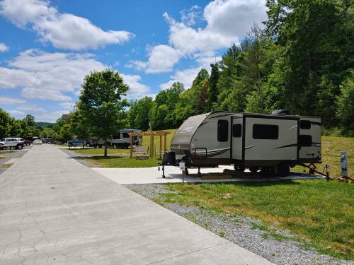 A scenic RV park with several campers, green trees, and a clear blue sky. Paved paths and grassy areas are visible.