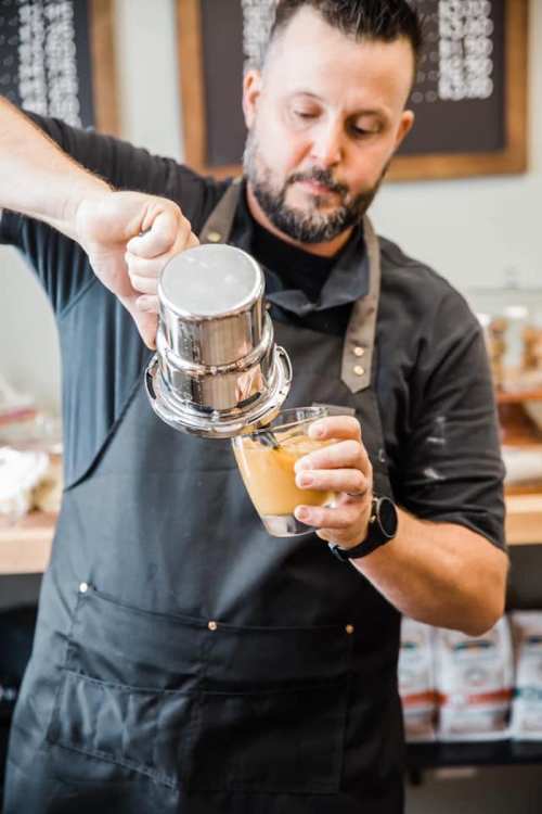 A barista pours coffee from a metal filter into a glass filled with ice, wearing an apron in a café setting.
