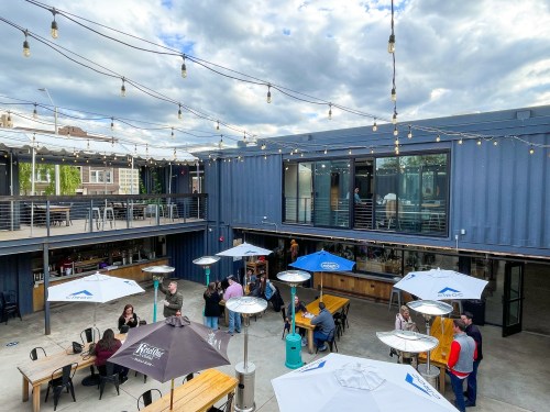 A lively outdoor space with string lights, people socializing, and tables under umbrellas at a container-style venue.