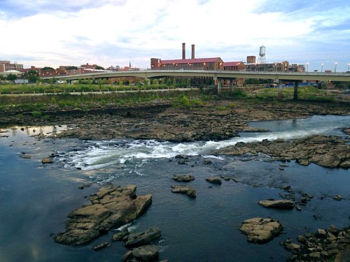 A river with rocky banks flows beneath a bridge, with a city skyline and industrial buildings in the background.