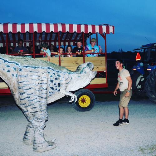 A person interacts with a dinosaur costume while a group of kids watches from a wagon in the background at dusk.