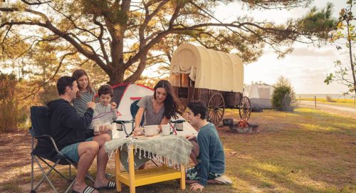 A family enjoys a picnic outdoors near vintage wagons, surrounded by trees and a peaceful landscape.