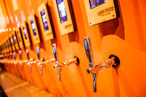 A row of shiny beer taps mounted on an orange wall, each with a digital display above.