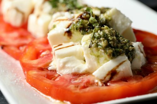 Fresh tomato slices topped with creamy feta cheese and a drizzle of pesto, served on a white plate.