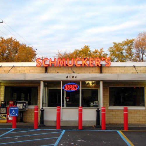 A storefront with the sign "Schmucker's" and an "OPEN" sign, featuring red poles and autumn trees in the background.