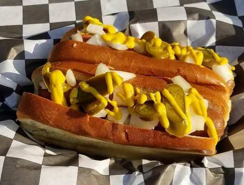 A hot dog topped with mustard, diced onions, and pickles, served in a checkered paper wrapper.