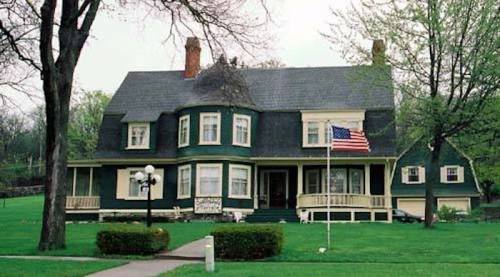A large green Victorian-style house with a porch, surrounded by trees and a lawn, featuring an American flag.
