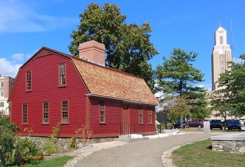 A red historic house with a shingled roof, surrounded by trees and a pathway, with a tall building in the background.