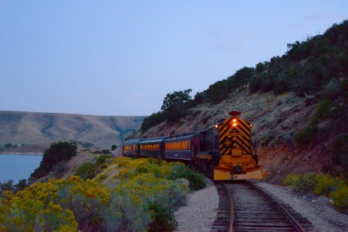 A train with glowing lights travels along a scenic track beside a lake, surrounded by hills and wildflowers at dusk.