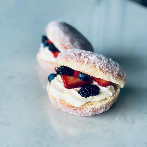 Two sugar-dusted doughnuts filled with cream and topped with strawberries and blueberries on a marble surface.