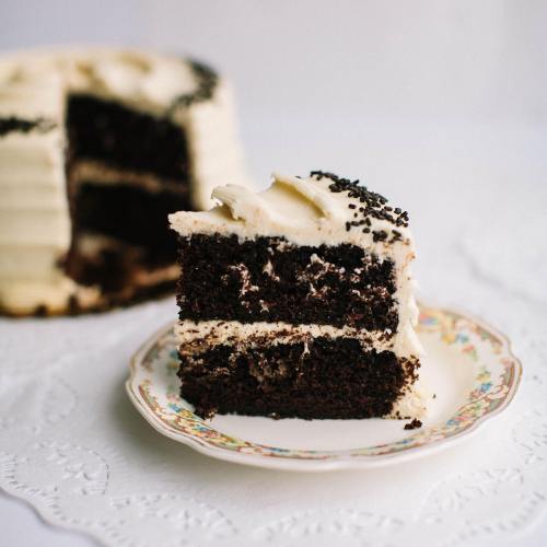 A slice of chocolate cake with creamy frosting on a decorative plate, with the whole cake in the background.