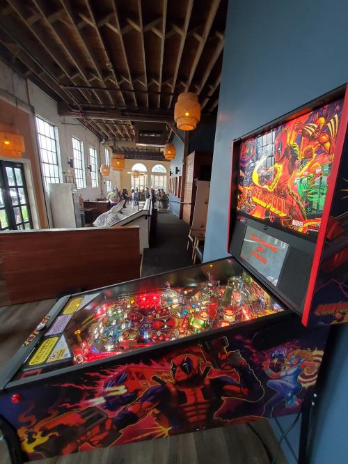 A colorful pinball machine featuring superhero graphics, set in a spacious, well-lit room with wooden decor.