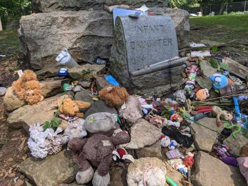 A gravestone marked "Infant Daughter" surrounded by various toys and flowers, creating a poignant memorial scene.