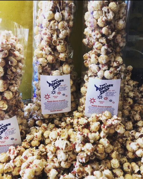 Bags of maple bacon popcorn stacked among loose popcorn, featuring colorful packaging and a festive design.