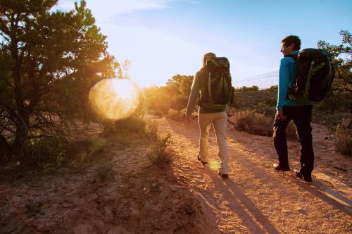 Two hikers with backpacks walk along a sandy trail at sunset, surrounded by shrubs and trees.