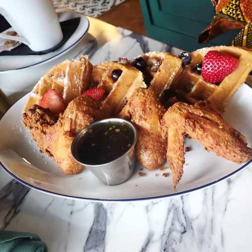 A plate of waffles topped with strawberries and blueberries, served with fried chicken and a small cup of syrup.