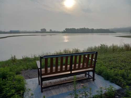 A wooden bench facing a calm lake at sunrise, surrounded by greenery and misty hills in the background.