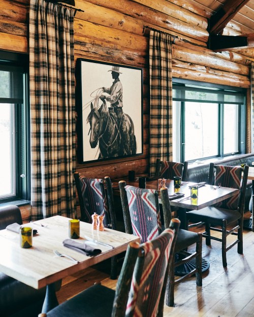 Cozy restaurant interior with wooden walls, plaid curtains, and a cowboy-themed artwork above rustic tables and chairs.
