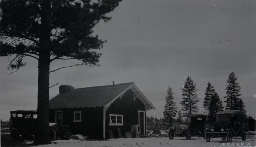 A vintage black-and-white photo of a small cabin surrounded by trees and two classic cars parked nearby.