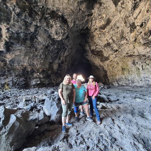 Four people stand inside a large cave, surrounded by rocky walls and a bright light at the cave's entrance.
