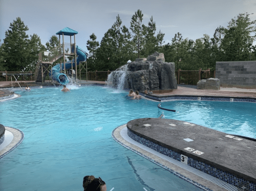 A pool area with a water slide, rock features, and people enjoying the water under a clear sky.