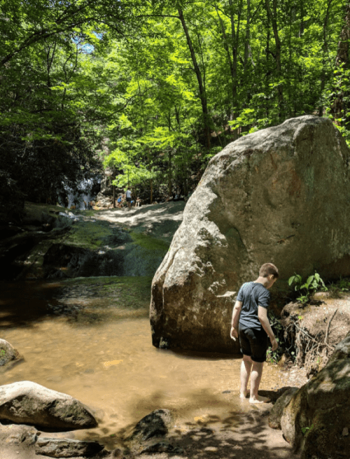 A boy stands by a shallow stream in a lush forest, with large rocks and people in the background near a waterfall.