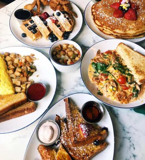A spread of breakfast dishes including waffles, pancakes, omelette, French toast, and potatoes on a marble table.
