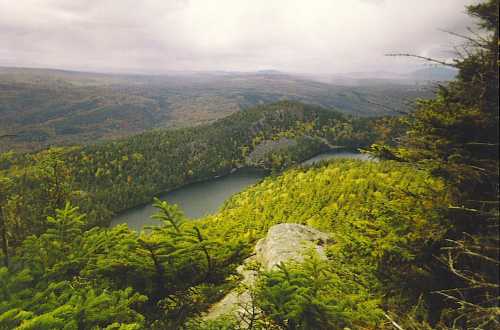 A scenic view of a lush green landscape with a winding lake surrounded by hills and trees under a cloudy sky.