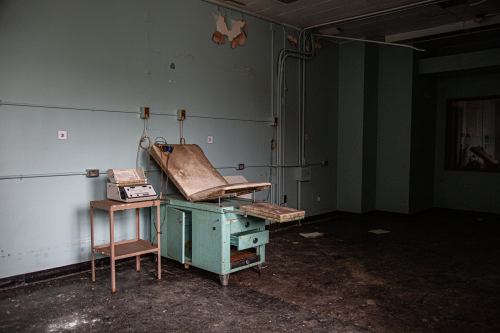 Abandoned room with a dusty examination table and old medical equipment against a peeling green wall.