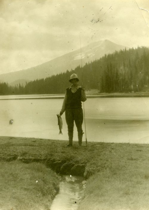 A person stands by a lake, holding a fish and a fishing rod, with mountains and trees in the background.