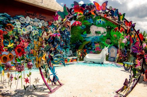 A vibrant outdoor art installation featuring colorful flowers, butterflies, and a white unicorn statue.