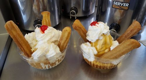 Two dessert cups with whipped cream, cherries, and churros, one topped with caramel and the other with a fruit topping.