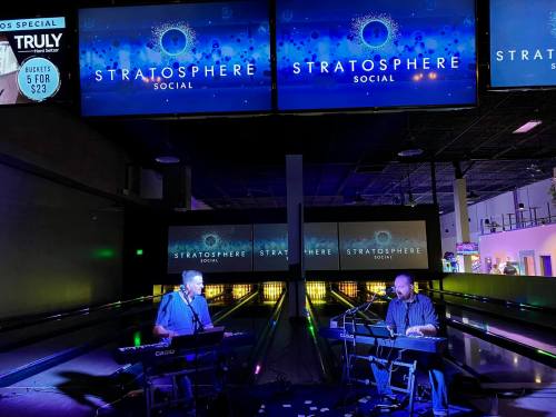 Two musicians perform on keyboards in a vibrant venue with large screens displaying "Stratosphere Social."