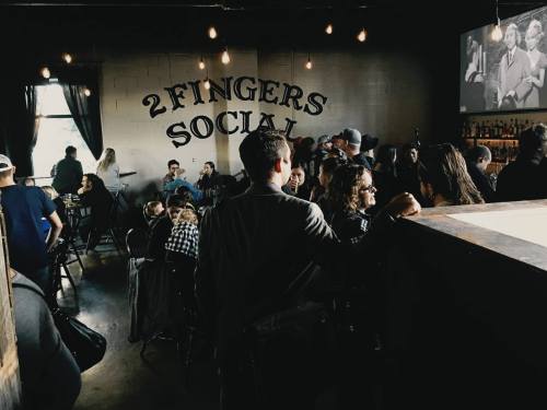 A lively bar scene at "2 Fingers Social," with patrons socializing and a vintage film playing on the wall.