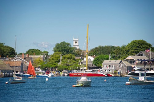 A scenic harbor with sailboats, a red boat, and charming buildings under a clear blue sky.