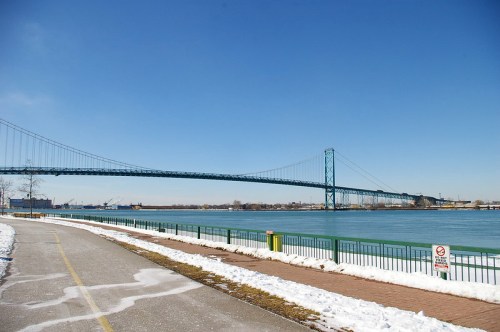 A wide view of a bridge spanning a river, with a clear blue sky and snow along the riverbank.