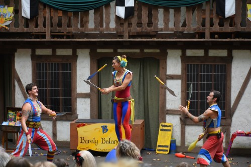 Three performers in colorful costumes juggle swords and entertain an audience in a lively outdoor setting.