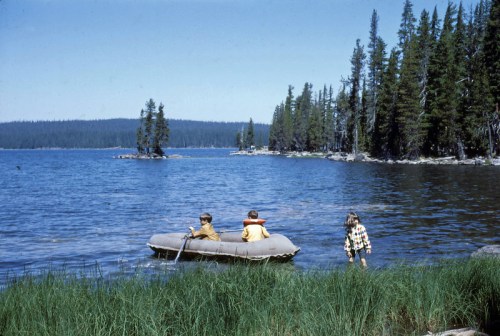 Two children in a small boat on a lake, with a girl walking along the shore and trees in the background.