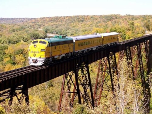 A vintage train crosses a tall trestle bridge surrounded by colorful autumn foliage.