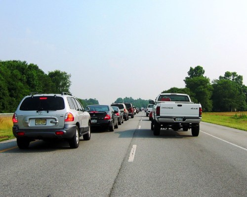 Traffic jam on a highway with several cars lined up, surrounded by green trees and a clear sky.