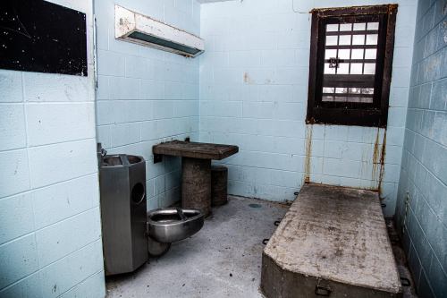 A stark, empty prison cell with a concrete bed, table, and a small barred window. Walls are painted light blue.