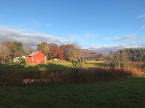 A red barn sits in a grassy field surrounded by trees and mountains under a clear blue sky.