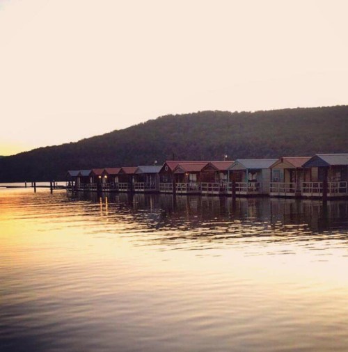 Colorful boathouses line a calm lake at sunset, reflecting on the water with a wooded hill in the background.