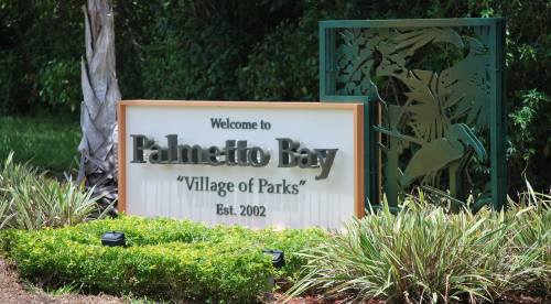 Welcome sign for Palmetto Bay, "Village of Parks," established in 2002, surrounded by greenery.